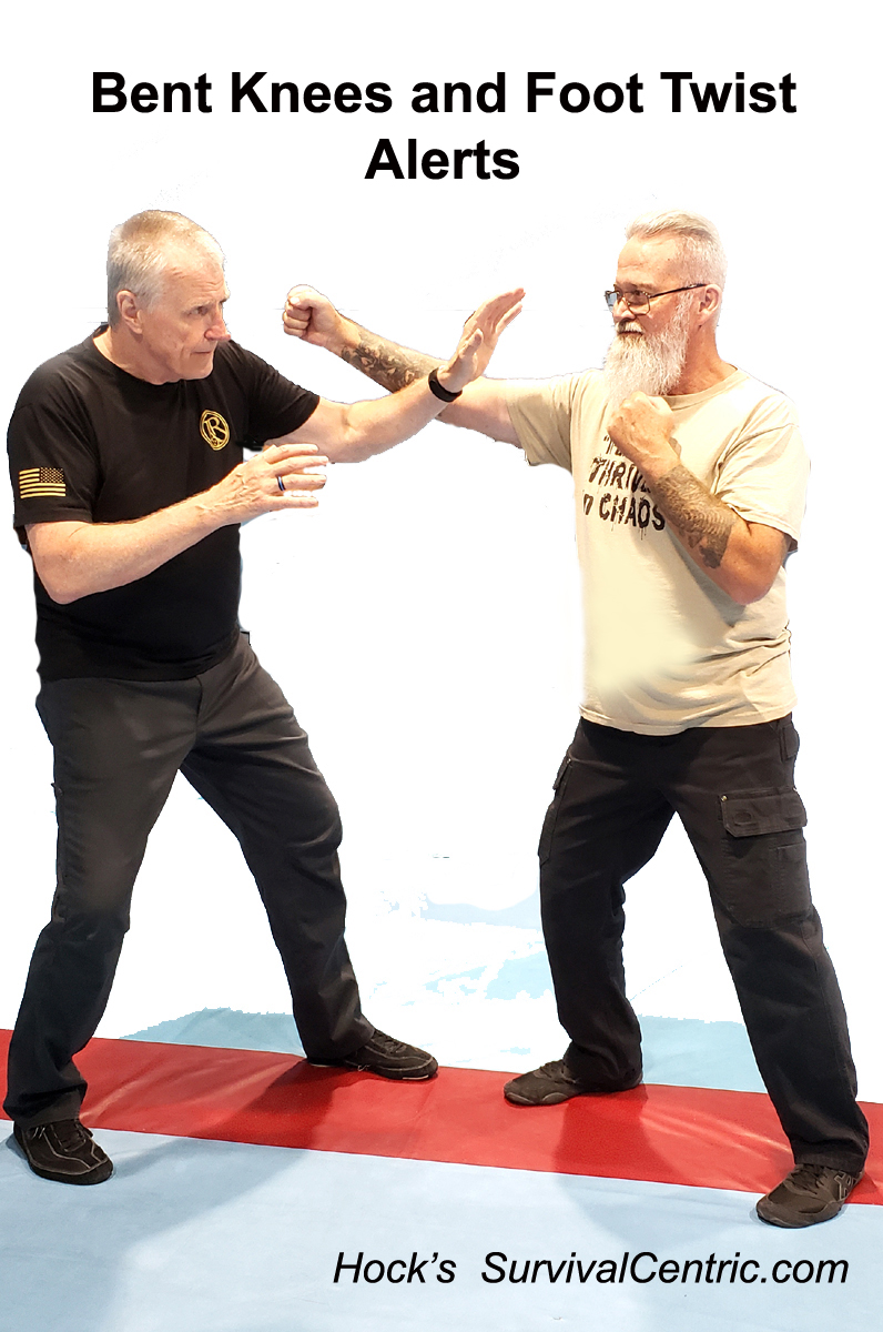 Two men on a padded mat demonstrate a bent-knee, foot-twist self-defense drill; one blocks with an extended forearm as the other presses forward.